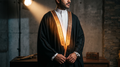 Man wearing a black ceremonial bisht with intricate gold-tone front borders over a white thobe, under a warm studio spotlight against a stone wall.
