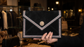 Black envelope clutch held by hand in a boutique, with triple metallic edging and a gold-tone sun-face medallion on the flap.