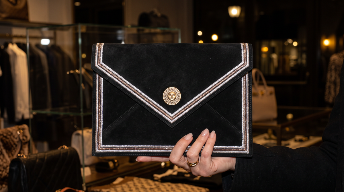Black envelope clutch held by hand in a boutique, with triple metallic edging and a gold-tone sun-face medallion on the flap.