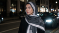Woman wearing a draped charcoal hijab with silver-grey geometric borders and a black coat on a city street at night.