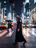 Man wearing a black thobe with white shoulder and sleeve detailing on a neon-lit city crossing at night.