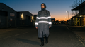 Woman wearing a long grey utility overcoat with bright bands across the torso and sleeves on a street at dusk.