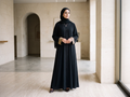 Woman wearing a black full-length abaya with gold-trimmed cuffs in a minimalist interior.